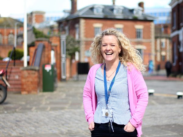 woman with blonde curly hair stands outside a building. She is smiling in a pink cardigan and blue top.