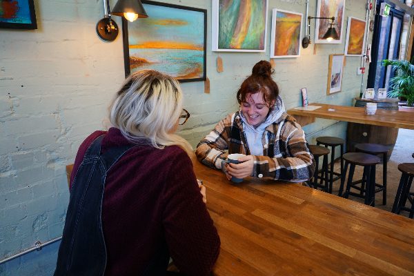 two young women sat across from one another talking and having coffee. one girl who is facing towards the camera is smiling