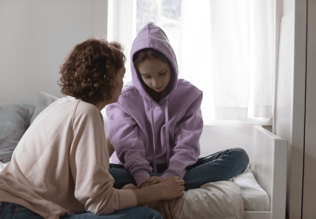 teenager sat cross legged in a purple hoody with the hood up looking down. A mother figure is holding her hands offering support.
