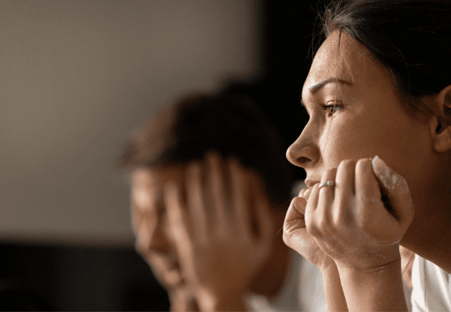 Man in the back ground with head in his hands. Woman in the foreground with chin resting in her hands looking concerned.